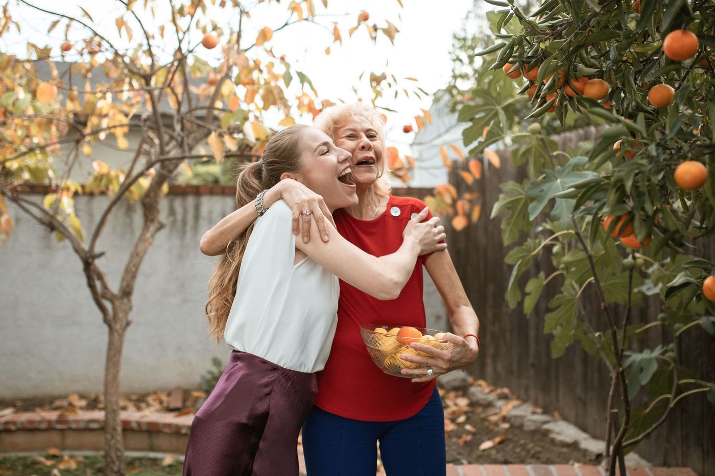 An older woman holding a fruit bowl and a younger woman laughing in a warm embrace in a backyard