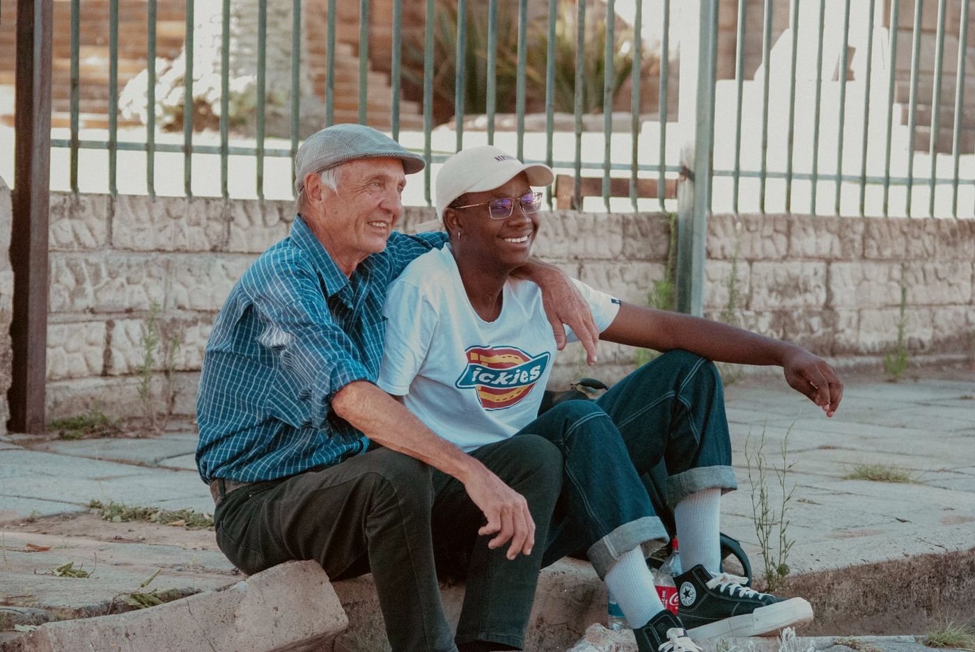 An older man in a flat cap with his arm around a smiling younger adult, seated together outdoors