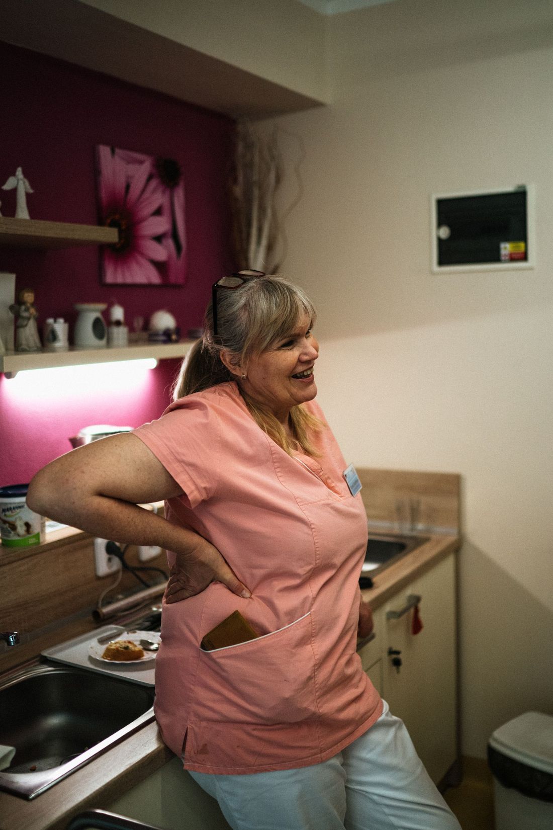 Woman in a pink uniform top laughing in a small kitchen