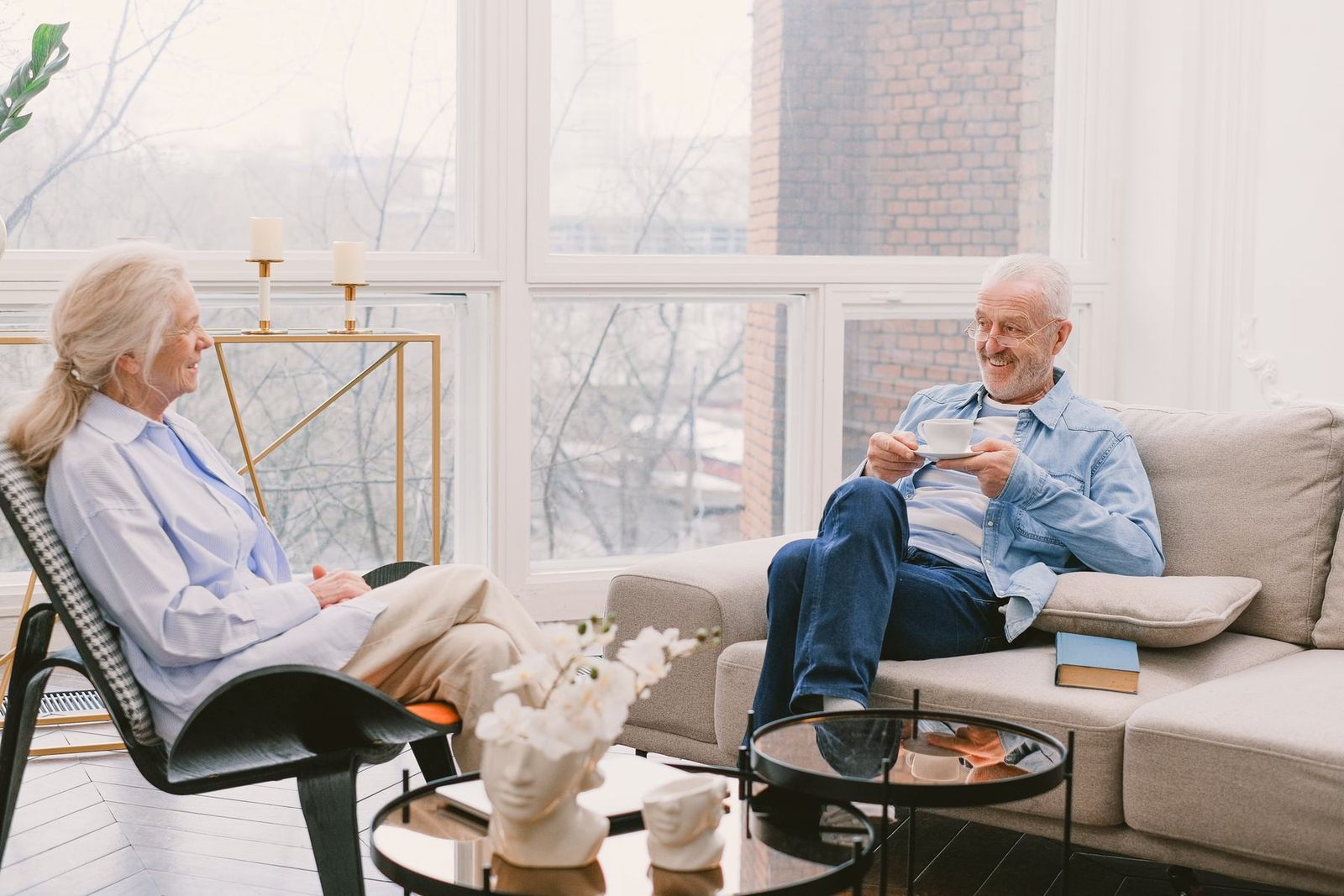 Two gray-haired people seated in a bright living room, laughing together over coffee