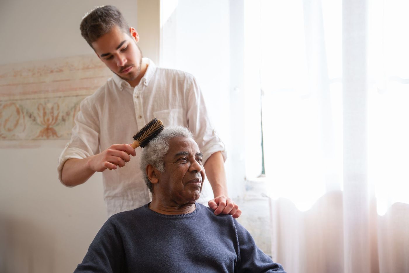 A younger man gently brushing a seated older man's hair beside a bright window